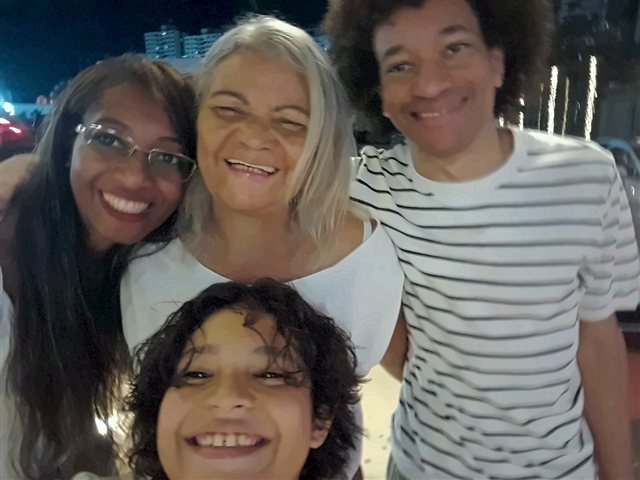 A Célia sorridente entre seus dois filhos adultos, com o neto João na frente